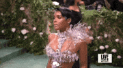 Met Gala 2024 gif. Slow motion clip panning down Janelle Monae's dress featuring a mostly see-through dress accented with holographic disks across her chest and down through the skirt. She wears a bursting silver poinsettia-like design at the top of one shoulder.