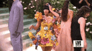 Met Gala 2024 gif. Slow motion shot of Nicki Minaj holding one hand to her head and looking up to her side in a playful pose. She's wearing a yellow Marni mini dress with fringe detailing, a low v-neckline, and 3D multicolored metal flowers sprouting in all directions around the dress. The skirt flares out and comes to her upper thigh as she dangles a tiny matching yellow purse.