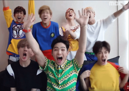 Celebrity gif. Members of GOT7, three sitting on a couch and four standing behind them, cheer and applaud, waving their hands, kicking the air, clapping and yelling.