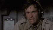 Video gif. Sweat pours off a stressed airplane pilots head as he wipes a hand across his wet brow. 