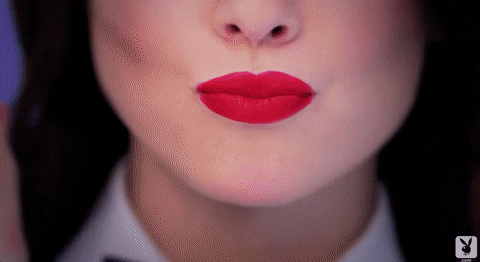 Video gif. We see a shot of a woman's nose and cherry red lips. She sends us a kiss before breaking out into a smile.