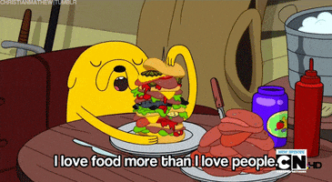 Cartoon gif. Jake from Adventure Time sits at a table in his tree house, holding onto a tall sandwich on his plate. His eyes are closed as he states, "I love food more than I love people."