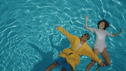 Music Video gif. From K.I.D.'s video for Errors, man floats on his back in a pool, wearing yellow goggles and a robe, next to a woman floating on her back, dressed in white.