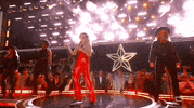TV gif. Country singer Lainey Wilson performing live on stage at the 2024 ACM Awards in bright red pants. She sings passionately into a microphone.