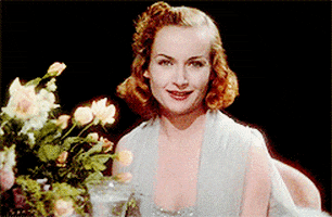 Celebrity gif. Vintage shot of Carole Lombard as Hazel in Nothing Sacred wearing a silver gown. She's sitting at a table with a flowers and lifts a crystal glass up at us shyly as she toasts us and takes a sip.