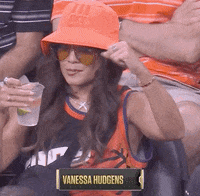 Celebrity gif. Vanessa Hudgens is at the 2021 NBA Finals and she wears a bucket hat and a jersey while holding a drink. She puts her hand in the air and does a stank face as she starts dancing along to the music.