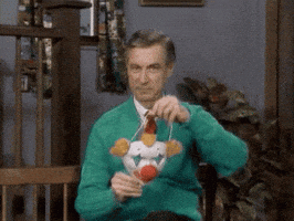 TV gif. Fred Rogers as Mr. Rogers puts on a clown mask with tufts of curly orange hair.