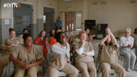 TV gif. Characters on Orange is the New Black sit and cheer on Taylor Schilling as Piper Chapman and Laura Prepon as Alex Vause as they stand in front of them, nervously smiling in front of them.