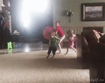 Video gif. The oldest of three kids throws a giant rubber ball at his siblings. The ball hits his brother, knocking him over, then bounces off a chair and knocks over the sister.