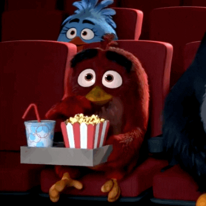 Cartoon gif. Red from the Angry Birds sits in a movie theater with a box that holds a soda and popcorn on his lap. He happily stares at the big screen as he shovels popcorn into his mouth.  