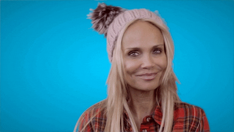 Video gif. Kristin Chenoweth wears a beanie with a pom-pom on top and looks at us as she smiles and gives a little wave. 
