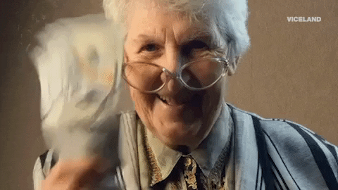 Video gif. A grinning old woman shakes a handful of one hundred dollar bills.