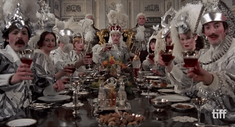 Movie gif. A scene from Tommy shows a long table full of food served on silver platters is surrounded by medieval lords and ladies and headed by a kingly looking man at the end. All the people wear monochromatic silver clothes as they look at us and toast with glasses of red wine.