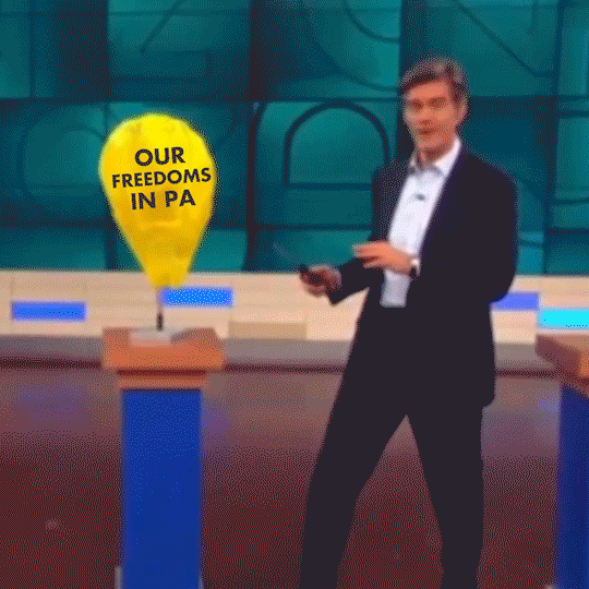 Video gif. Dr. Oz holds a lighter to a yellow balloon labeled “Our freedoms in PA,” dramatically raising his hands in the air as it explodes into flames.