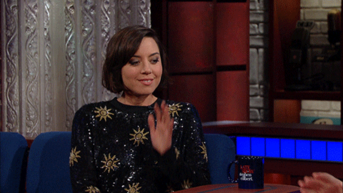 Celebrity gif. Aubrey Plaza rests her elbow on The Late Show desk, cupping her chin in her palm and glancing to the side with a coy smile.
