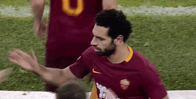 Sports gif. Mohamed Salah in an AS Roma football uniform shakes hands and hugs teammates on the field with a serious, stoic expression on his face.
