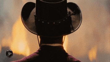 Movie gif. Jamie Foxx as Django in Django Unchained wears a cowboy hat and stares into a large fire, facing away from us. He then turns to face us, wearing circular sunglasses with a long cigar in his mouth, and grins with a slight head nod.