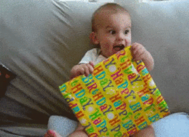 Video gif. A baby is holding a birthday present and looks ecstatic. The camera runs in on their face and they open their mouth in joy.