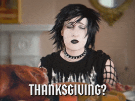 Video gif. Goth person dressed in all black with black makeup and hair pouts as they sit at a dining table for thanksgiving. Text, “Thanksgiving, thanks for nothing.”