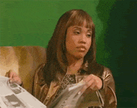 Video gif. Woman sits on a couch, holding a newspaper in her hands. She rolls her eyes at someone and holds the newspaper up to hide from whoever she’s annoyed with.