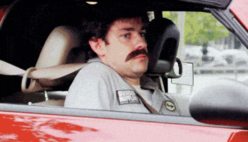 The Office Gif. John Krasinski as Jim Halpert sits in the passenger seat of a car. He wears a fake mustache and a Dunder Mifflin Warehouse uniform as if he’s in disguise. He looks out the window with a look of shame and slides the back of his seat down so he slowly disappears from view.