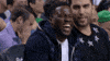 Sports gif. Kevin Hart sits courtside at a basketball game in a dark blue coat, laughing at a comment from the stubbly man next to him.