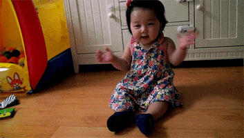 Video gif. Baby sits on wood floor, wearing a floral-print dress, smiling wide and clapping slowly.