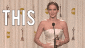 Celebrity gif. Jennifer Lawrence stands in front of a microphone with an Oscar award in her hand. She looks around with big eyes and gestures her arms out, then slapping her side. Text, “This.”
