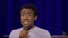 Celebrity gif. Donald Glover looks at the camera with a still grimacing expression. He tugs on the collar of his shirt and winces.