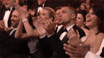 TV gif. A camera zooms out on a large crowd of celebrities sitting at an award show. The front row clap, sitting down, and laughing with wide smiles on their faces. The rows behind them stand up to give a standing ovation, cheering on the person on stage with immense enthusiasm. 