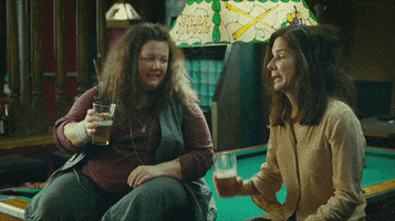 Movie gif. Melissa McCarthy Detective Shannon and Sandra Bullock as Special Agent Sarah. The two ladies are sitting on a pool table in a bar and they clink beer glasses but they underestimate their strength and the glasses shatter.