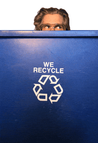 Video gif. A man peeks out from behind a blue recycling bin. We can only see half his face and he's scanning up and all around in a shifty or nervous way.