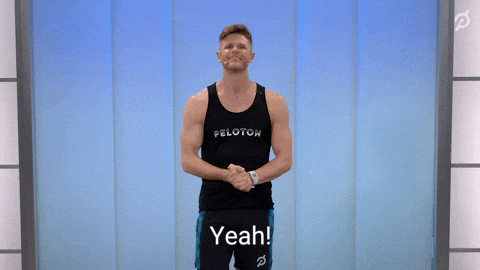 Sports gif. Andy Speer wears a Peloton tank as he smiles with his hands clasped at his waist and nods affirmatively.