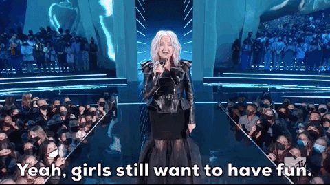 Celebrity gif. Cyndi Lauper performing at the 2021 VMAs. She is on stage and holds a microphone in her hand as she announces, "Yeah, girls still want to have fun," and the crowd around her cheers when she points at them.
