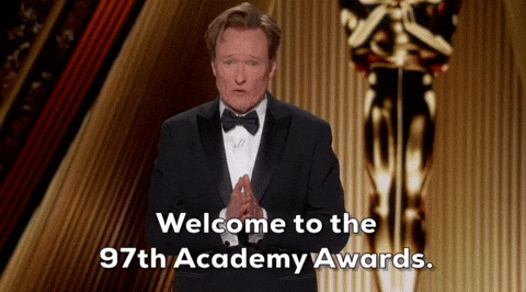 Conan O’Brien, wearing a tuxedo and looking earnest with his hands together, stands in front of a golden toned set with the Oscar statuette. Text below reads, “Welcome to the 97th Academy Awards.”