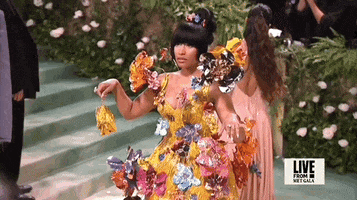 Met Gala 2024 gif. Slow motion closeup of Nicki Minaj walking forward in her yellow Marni mini dress with a low-cut v-neckline and 3D multicolored metal sprouting flowers that seem to sprout all around her and a stiff flared skirt. She holds up a tiny matching yellow purse.