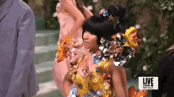 Met Gala 2024 gif. Closeup of the top of Nicki Minaj's yellow Marni dress with fringe detailing, a low-cut v-neckline, and 3D multicolored metal flowers that seem to sprout all around her. Her hair is styled in a bob ringed with a colorful floral crown and straight bangs.
