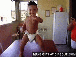 Video gif. Baby stands on a table dancing with one arm out, shaking their hips and turning from side to side.