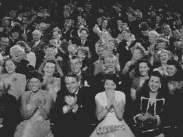 Video gif. Black and white footage shows a large, well dressed audience clapping and cheering excitedly with big smiles on every face.