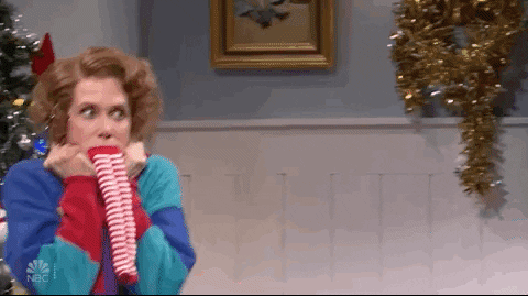 SNL gif. Kristen Wiig as Sue, wriggles next to a Christmas tree, hands clutching her turtleneck, stocking over her mouth, trying but failing to contain her exuberant excitement.