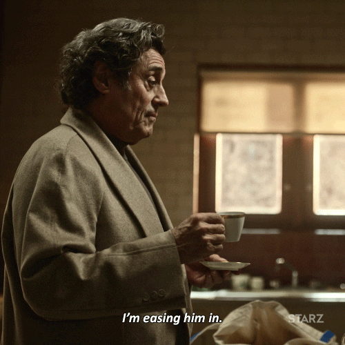 TV gif. Ian McShane as Mr. Wednesday on American Gods hods a small tea cup and saucer in his hands while standing in the kitchen. He looks down at someone as he says, “I’m easing him in.”