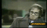 Video gif. A man looks at us with half circle glasses on his face. He has a big grin on his face while he makes an “A-ok” symbol with his hand. 