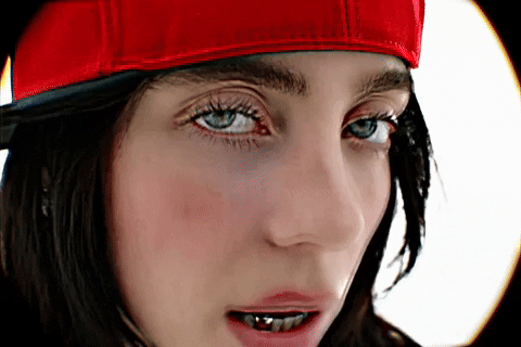 Music Video gif. From Billie Eilish's music video for "LUNCH", Billie is on a stark white background dressed in a red flat brim hat. She is looking into the camera while singing her song.
