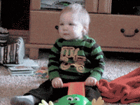Video gif. A baby sits in a living room holding a push toy, dancing and wiggling around, moving his shoulders. 