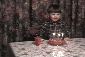 Video gif. Old home video of a child headbanging while blowing out three birthday candles on a cake.