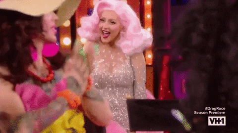 Reality TV gif. Christina Aguilera on RuPaul’s Drag Race wears a big pink wig and a completely bedazzled dress. She says “Hi” and grins widely. She holds up her fluffy boa scarf and shakes her hands to wave at the Drag Queens. 
