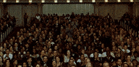 Movie gif. In a huge, fancy auditorium with multiple balconies, the entire audience begins to stand up and applaud.