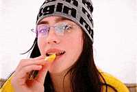 Music Video gif. From Billie Eilish's music video for "LUNCH", Billie is on a stark white background dressed in a bright yellow shirt and a black and grey knit hat and silver glasses. She is staring into the camera while eating what looks to be a french fry.
