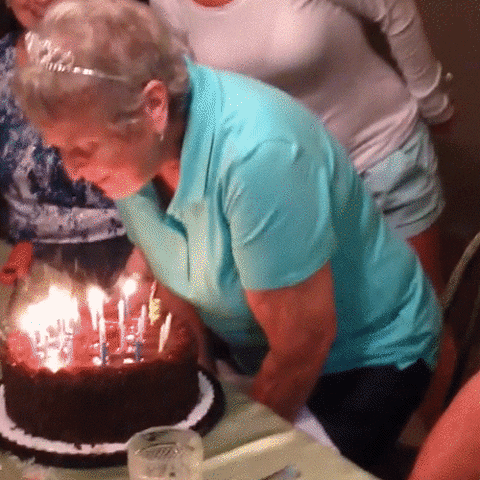 Video gif. Old woman blows out the candles on her birthday cake, sits back down, and then everybody around her sprays her with silly string.