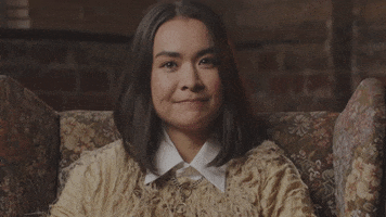 Celebrity gif. Mitski sits in a chair and gazes directly at us with a taut smile.